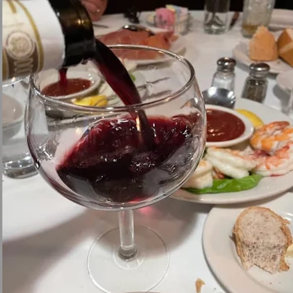 Red wine pouring into a glass, with shrimp cocktail and bread on a white tablecloth.