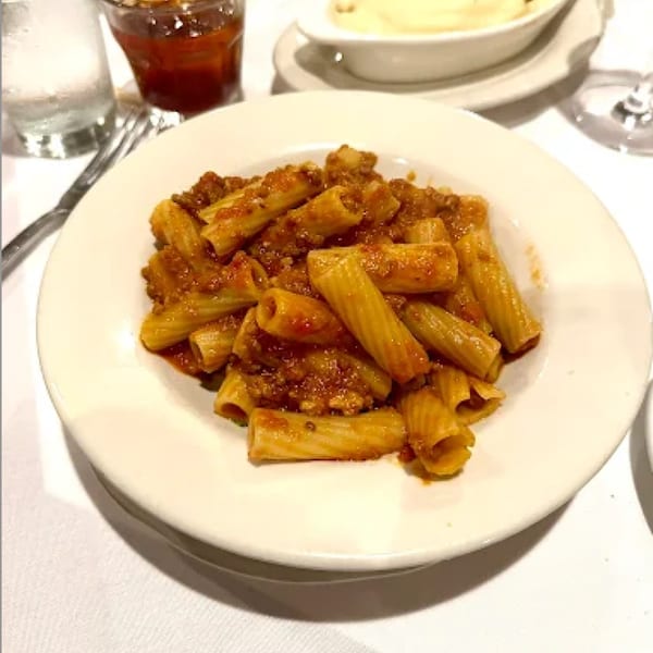A white plate of rigatoni pasta with a rich meat sauce, garnished with grated cheese.